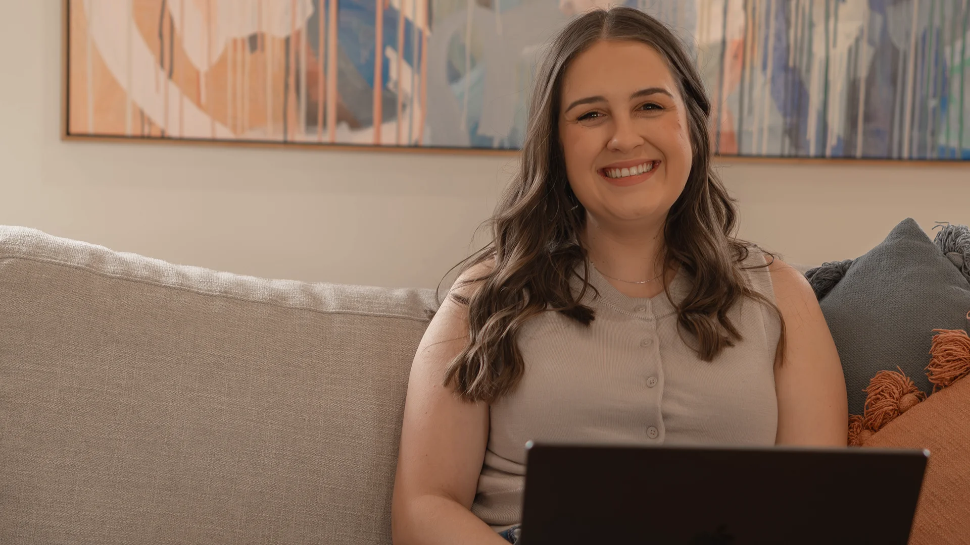 Elyse Williams of Elyse Williams Creative sitting on a couch on her laptop smiling at the camera with colourful painting in background