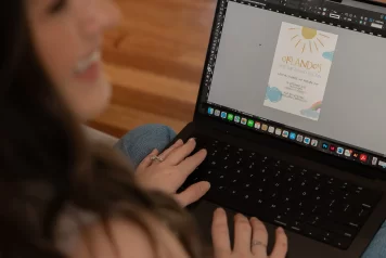 Elyse Williams Creative's laptop screen in focus showing a first birthday invitation being worked on in InDesign